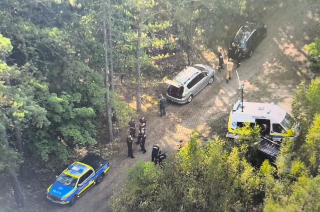 Grupa policjantów prowadzi działania w lesie, obok zaparkowanych samochodów.