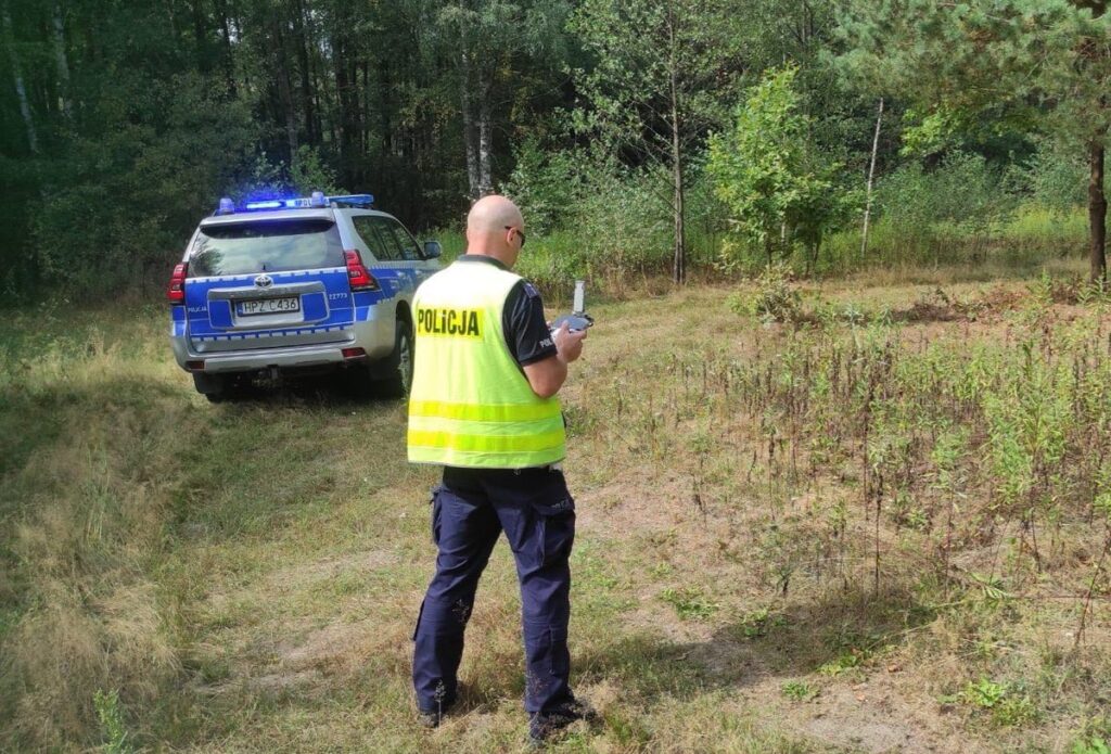 Policjant w żółtej kamizelce kontroluje teren leśny z ręcznym urządzeniem, obok zaparkowany radiowóz.