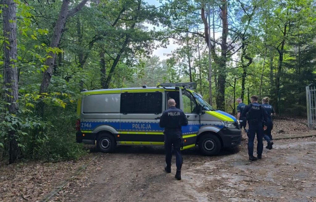 Grupa policjantów w mundurach podchodzi do radiowozu zaparkowanego w lesie.