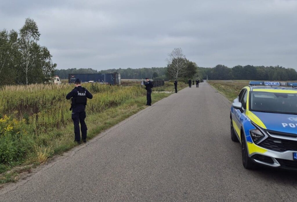 Policjanci przeszukują teren przy drodze, obok stoi radiowóz policyjny. W tle widoczna zarośnięta łąka.
