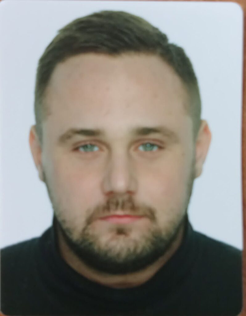 Mężczyzna z zarostem w czarnym golfie, patrzy prosto w obiektyw na tle białego tła.