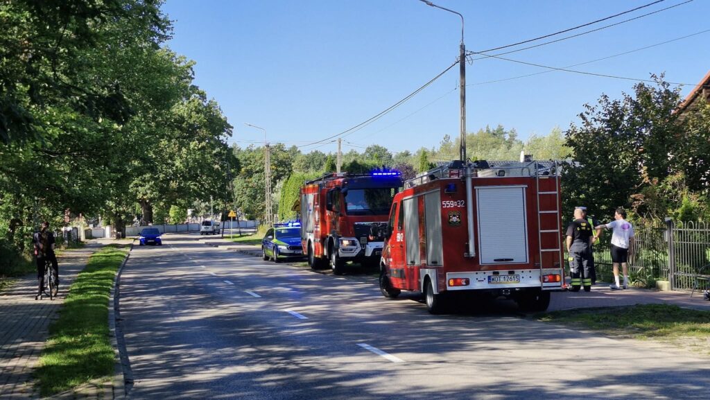 Dwa wozy strażackie oraz radiowóz policji zatrzymane na poboczu drogi, obok grupy osób rozmawiających w słoneczny dzień.