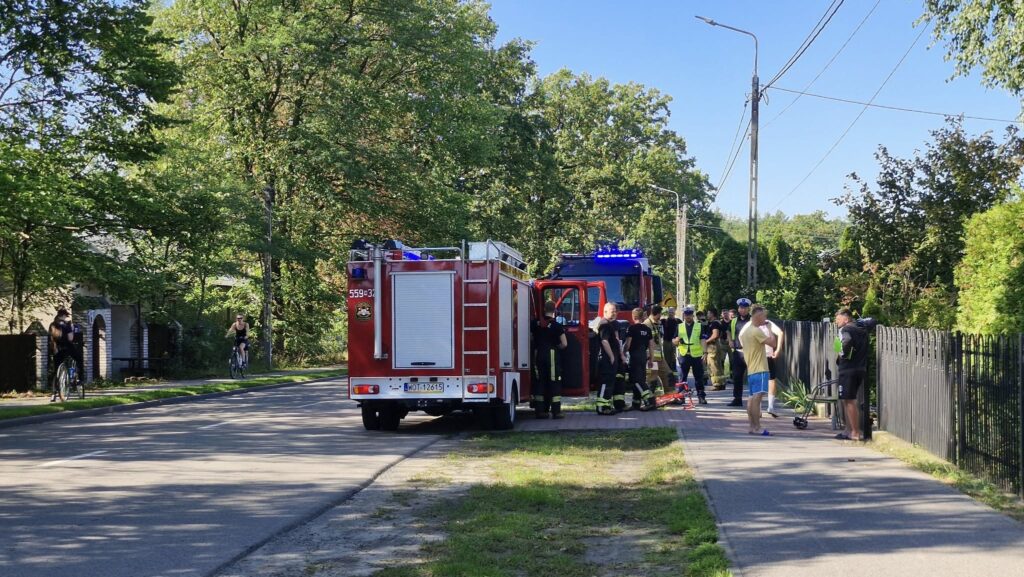 Zespół strażaków w mundurach omawia sytuację obok wozu strażackiego na ulicy, w otoczeniu zieleni.