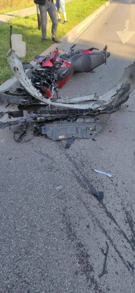 Zniszczony motocykl leżący na asfalcie, wokół widoczne fragmenty pojazdu i ślady oleju. W tle stoją dwie osoby.