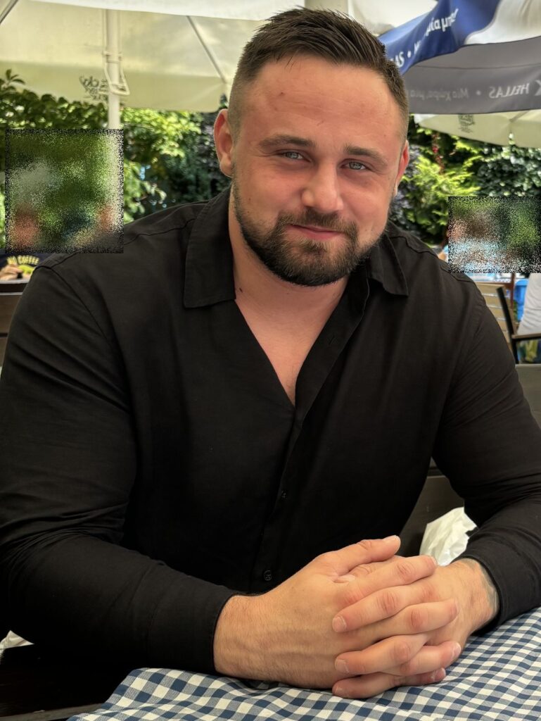 Mężczyzna w czarnej koszuli siedzący przy stole z niebiesko-białym obrusem w kawiarni na świeżym powietrzu.