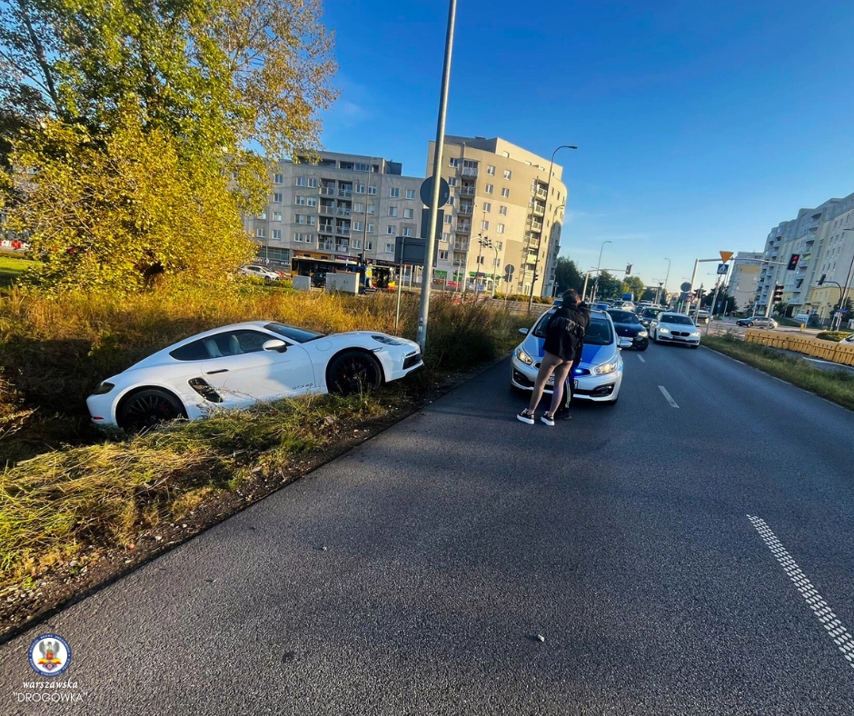 Biały sportowy samochód w rowie przy drodze, obok dwóch osób rozmawiających przy zatrzymanym pojeździe.