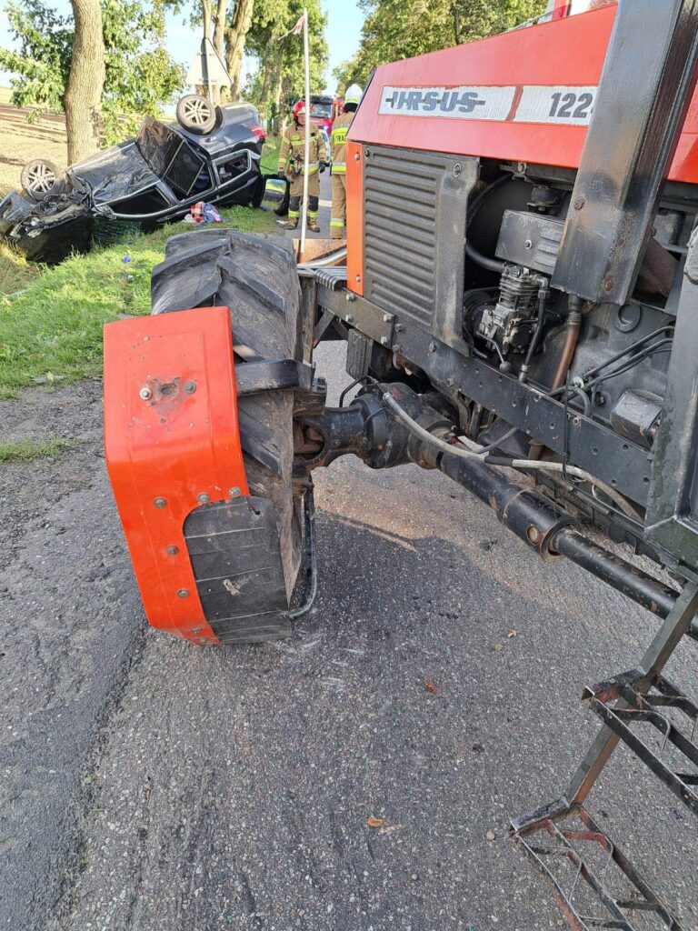 Traktor Ursus z uszkodzoną tylną oponą stoi na drodze, obok przewrócone auto osobowe oraz strażacy w mundurach.