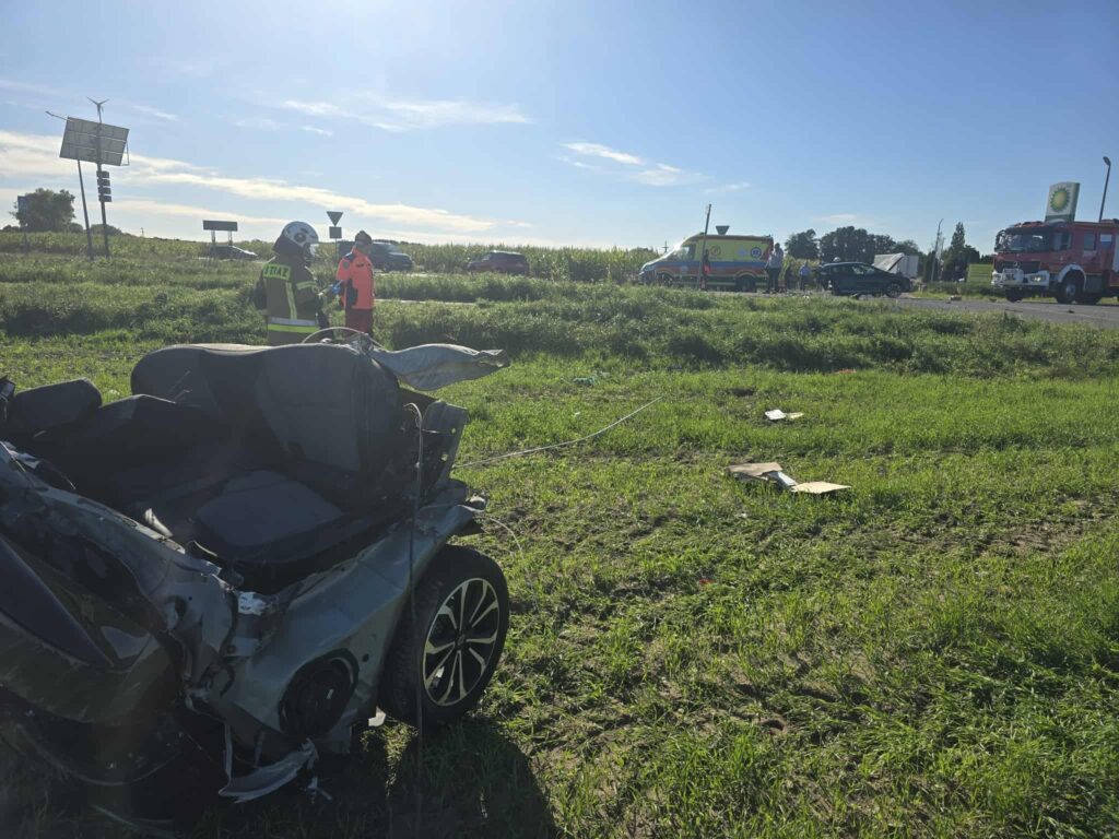 Zniszczony samochód stoi na poboczu drogi, obok pracują strażacy i ratownicy medyczni.
