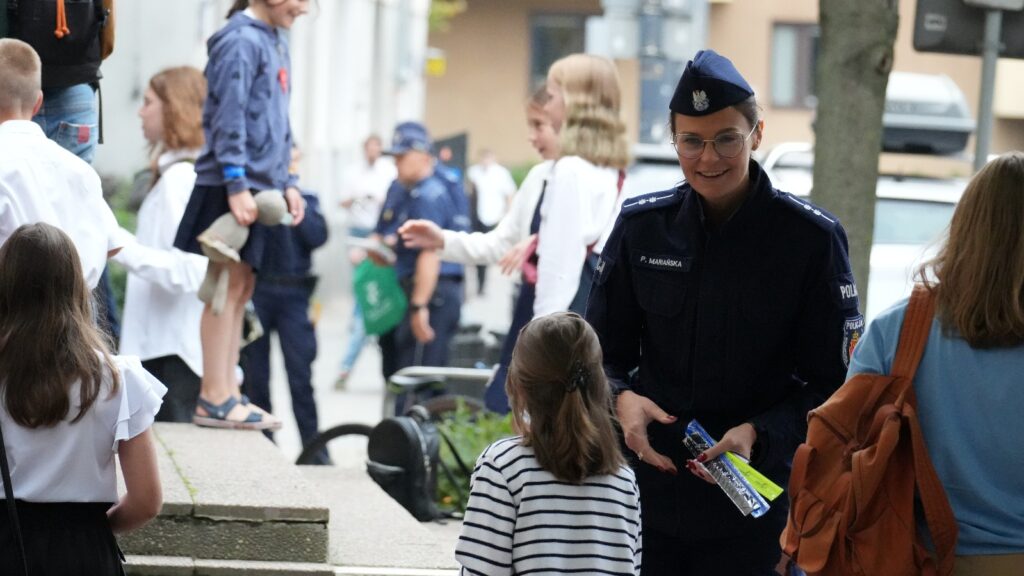 Policjantka rozmawia z dzieckiem na chodniku, w tle widać grupę uczniów i mundurowych.