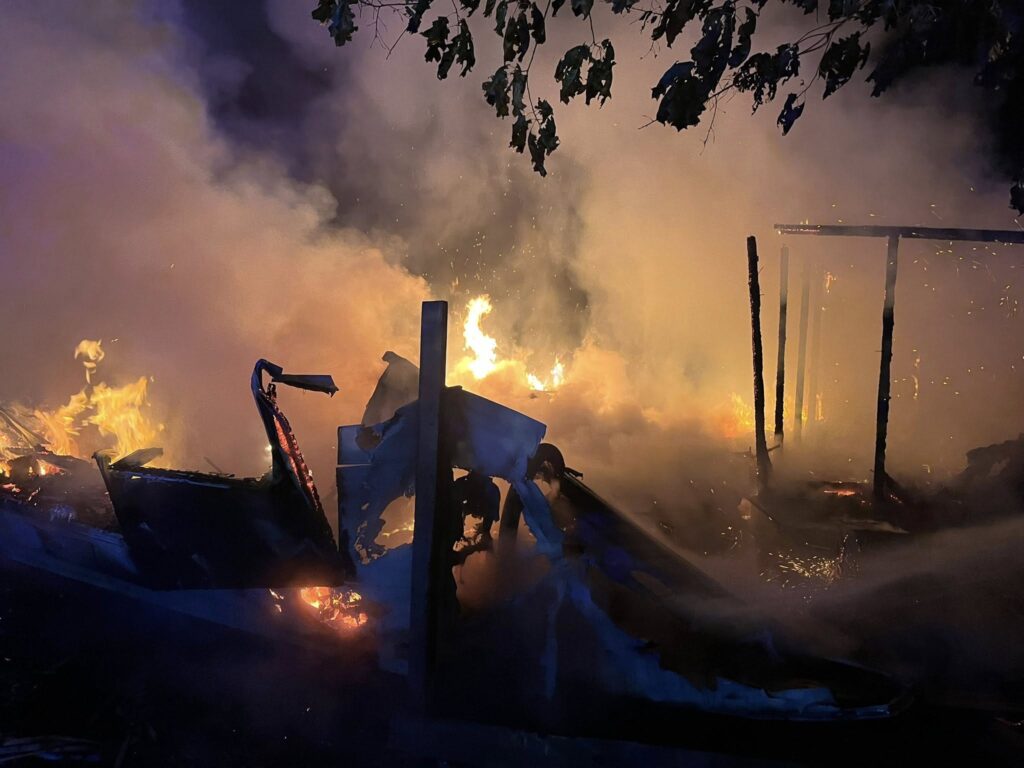 Płonąca konstrukcja wypełniona dymem, z widocznymi płomieniami i zgliszczami, otoczona ciemnym tłem.