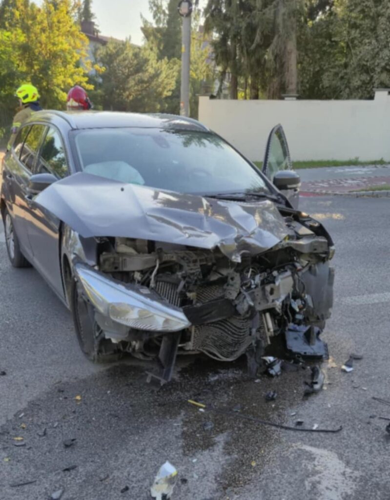 Uszkodzony samochód osobowy stoi na drodze, obok niego leżą fragmenty wraku oraz plama oleju.