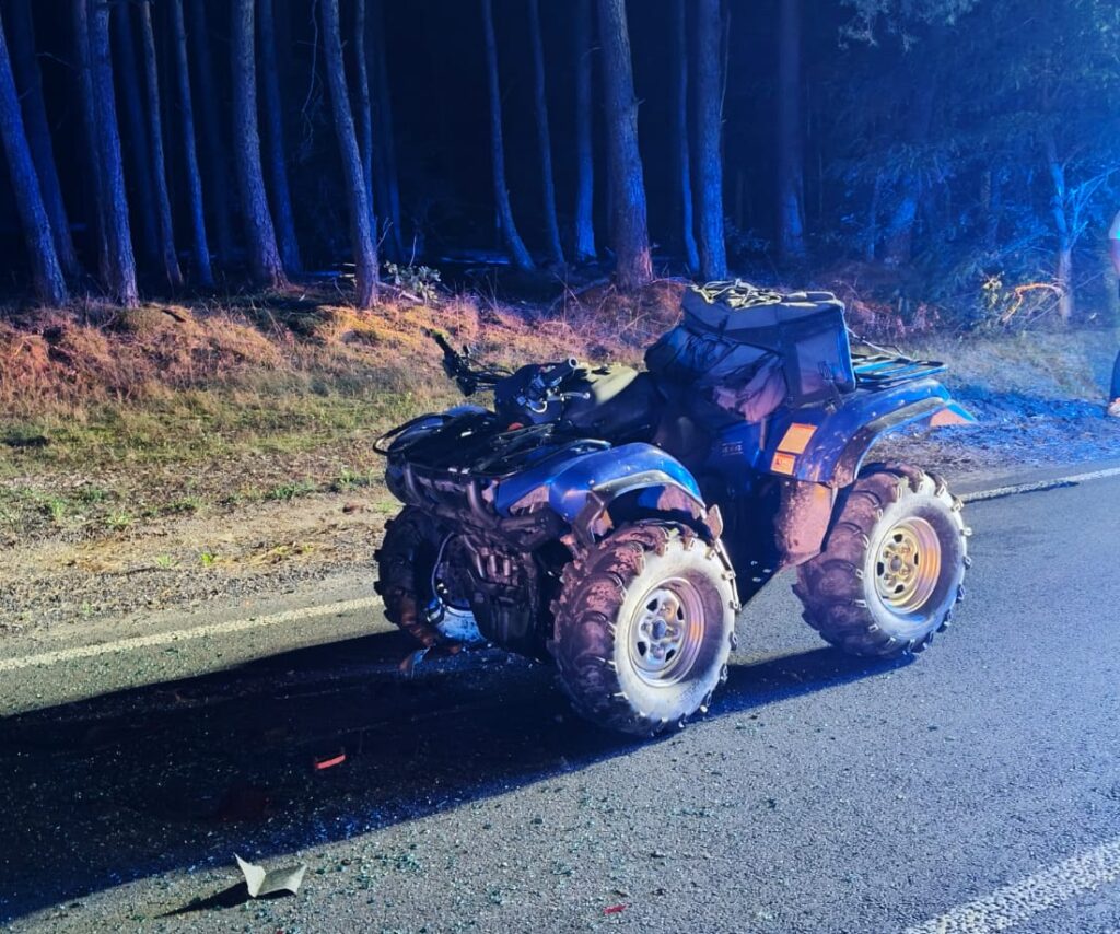 Quad leżący na drodze, otoczony rozbite szkło, w tle ciemny las i niebieskie światła policyjne.