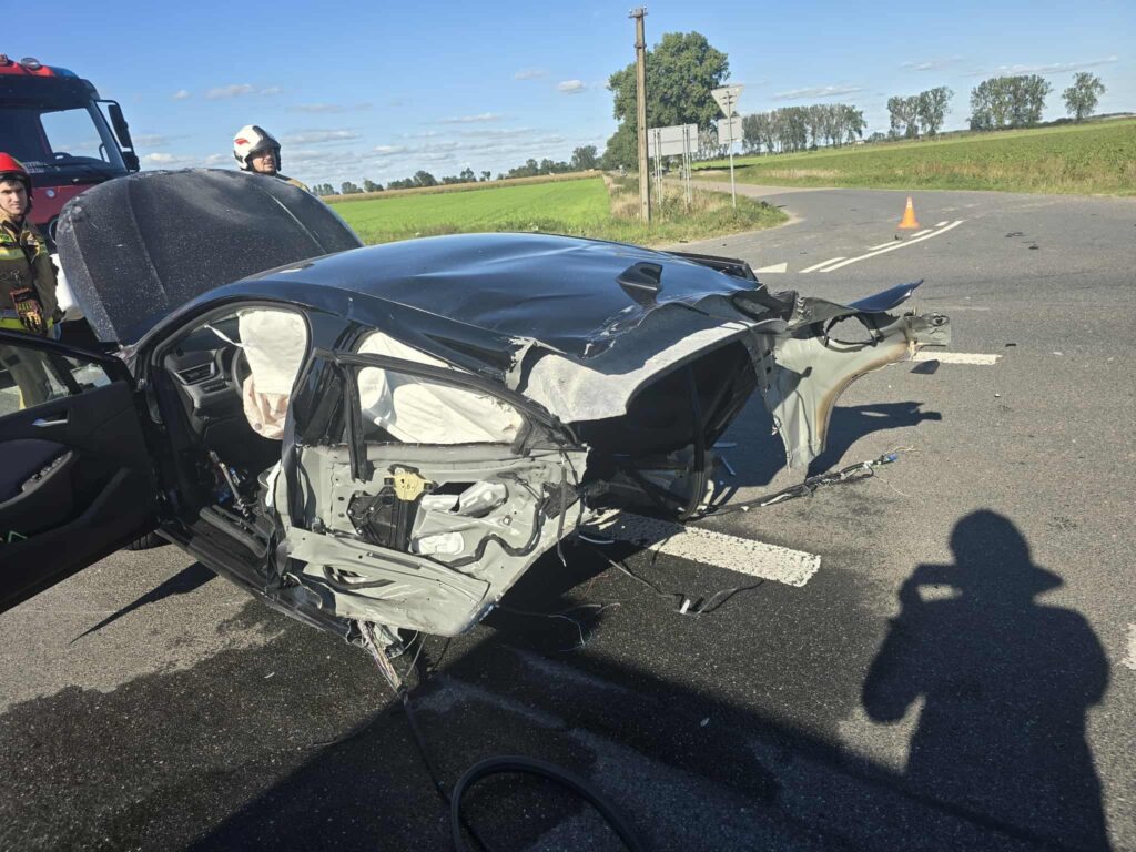 Rozbita osobówka z uszkodzoną stroną, obok stoją strażacy w hełmach, w tle zielone pola.