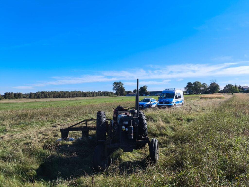 Traktor stojący na łące obok radiowozu i karetki, w tle rozległe pola i drzewa.