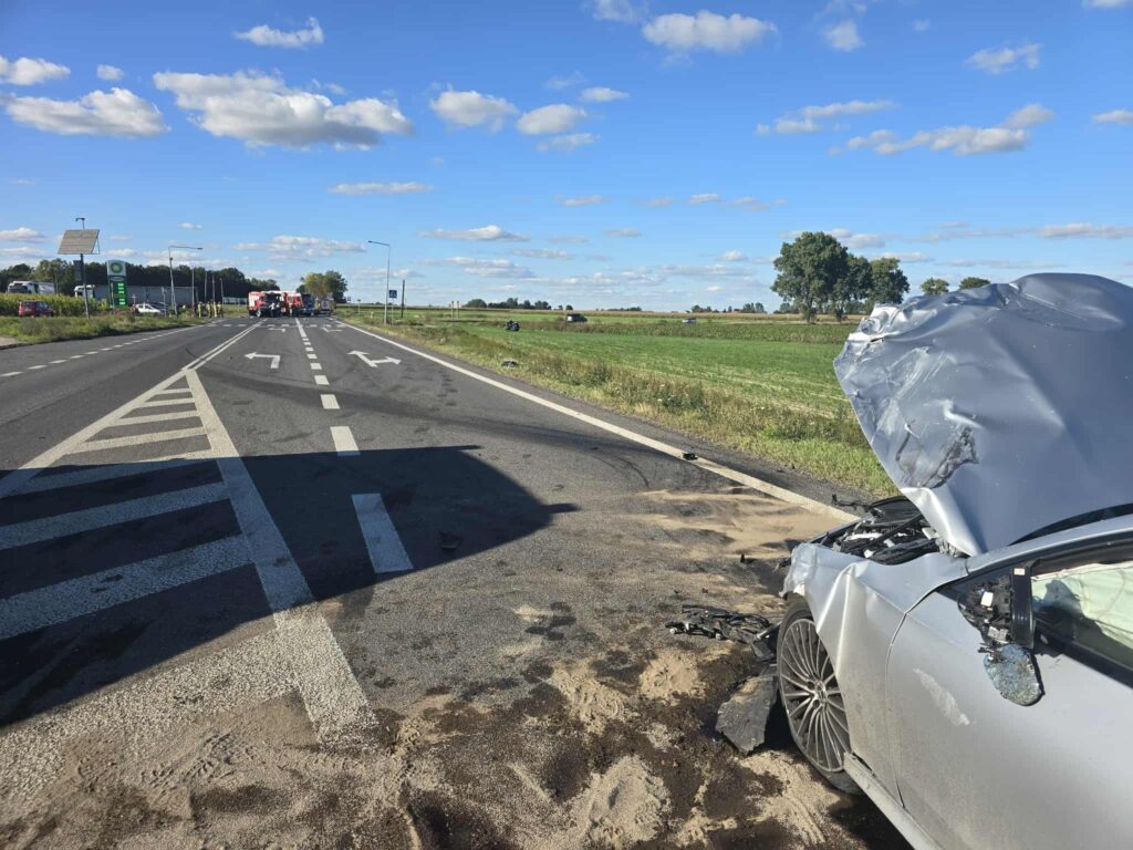 Uszkodzony samochód osobowy z otwartą maską leży na drodze, obok widać pojazdy służb ratunkowych. W tle rozciągają się pola.