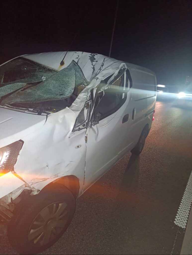 Uszkodzony biały samochód dostawczy z rozbitą szybą i wgniecioną karoserią stoi na drodze w nocy.