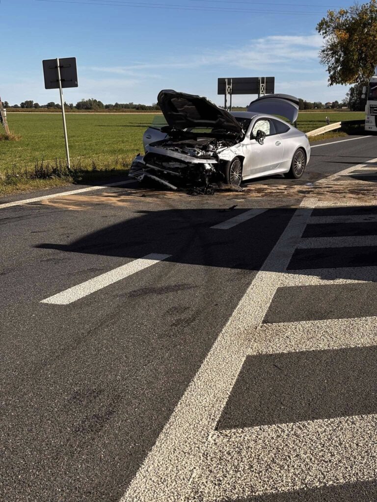 Zniszczony samochód osobowy z otwartą maską stoi na drodze, obok znaków drogowych i zielonego pola.