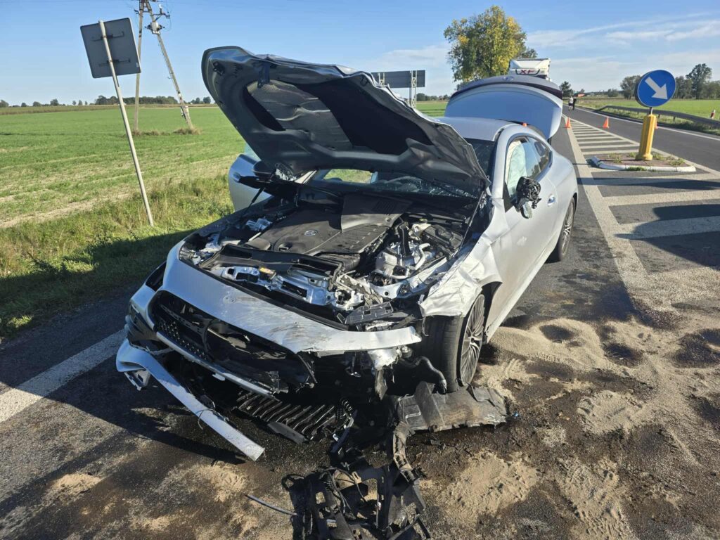 Uszkodzony samochód osobowy z otwartą maską stoi na drodze, obok znaku drogowym i zielonym polem.