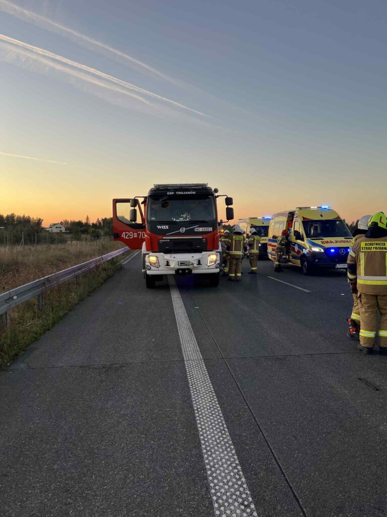 Strażacy pracują przy wypadku drogowym, obok wozu strażackiego i ambulansów na drodze w czasie zachodu słońca.