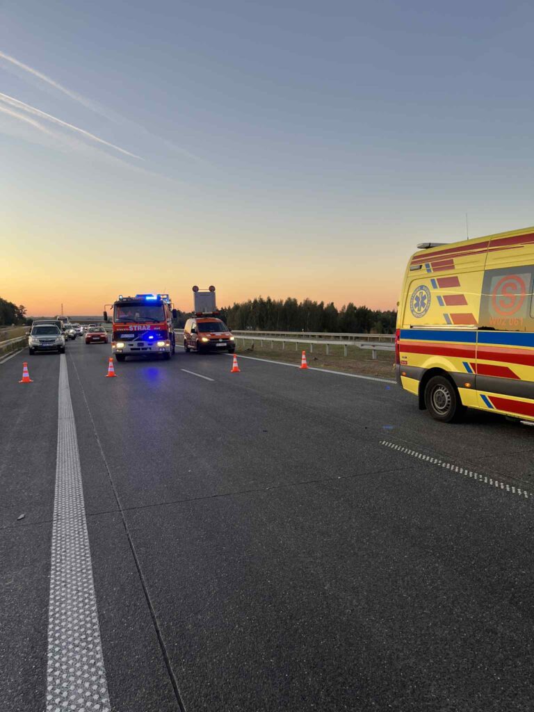 Na drodze stoi kilka pojazdów ratunkowych z włączonymi sygnałami świetlnymi, a w tle widać zachód słońca.
