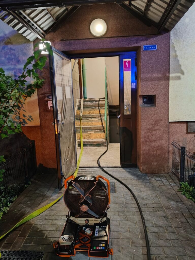 Wentylator przemysłowy zasilany elektrycznie stoi na chodniku przed otwartymi drzwiami budynku, prowadzącymi na schody.