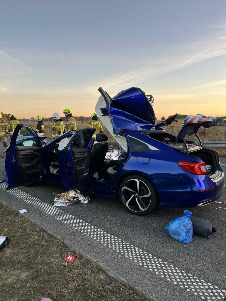 Nieoznakowany niebieski samochód osobowy z rozciętą karoserią stoi na poboczu drogi, a obok pracują strażacy w kaskach.