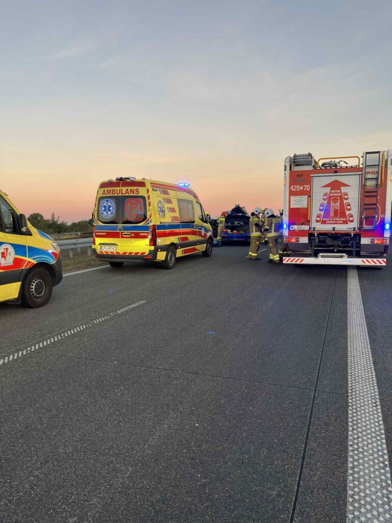 Zespół ratunkowy z ambulansami i wozem strażackim interweniuje na drodze w trakcie zachodu słońca.