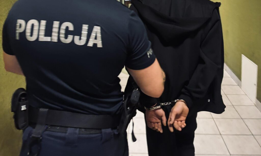 Policjant prowadzi aresztowanego mężczyznę w kajdankach korytarzem w budynku.