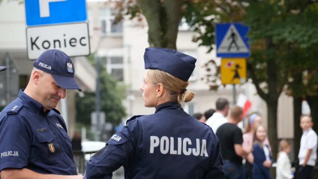 Dwóch policjantów rozmawia na ulicy, w tle widoczne znaki drogowe oraz grupka ludzi.