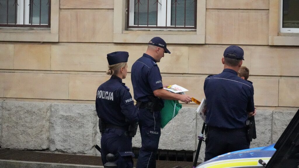 Policjanci rozmawiają z dzieckiem, trzymając w rękach materiały informacyjne, obok budynku z oknami.