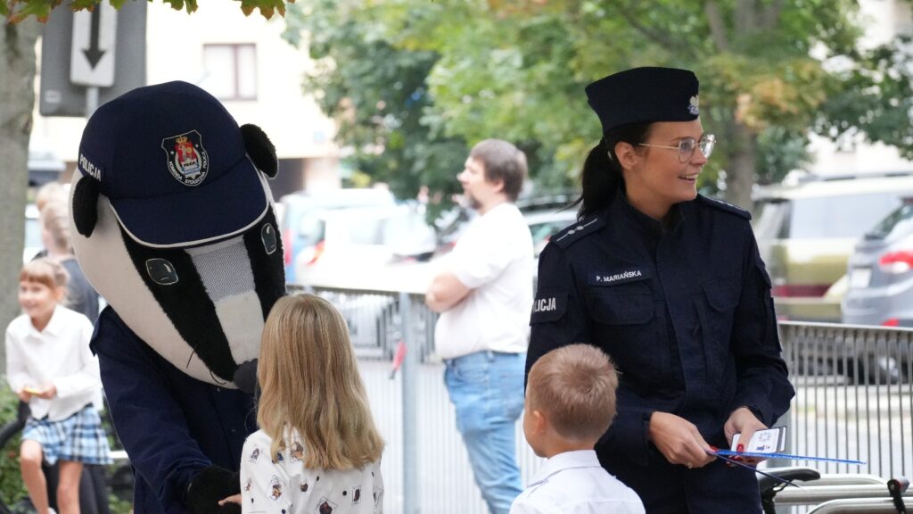 Mężczyzna w przebraniu borsuka rozmawia z dziećmi, obok stoi policjantka w mundurze. W tle widać ruch uliczny.