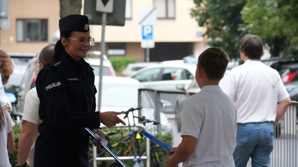 Policjantka rozmawia z chłopcem, trzymając w ręku niebieską pałkę, w tle widoczne samochody i osoby przechodzące obok.