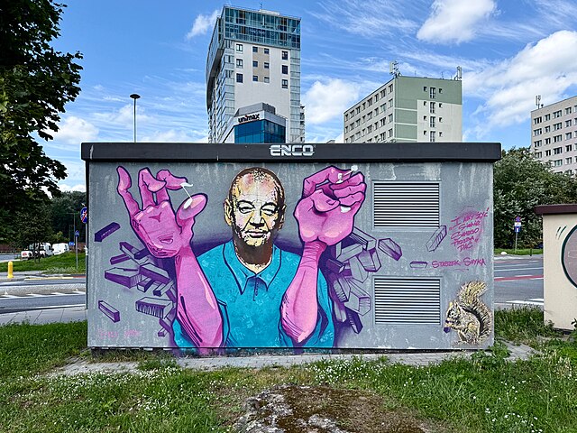 Mural przedstawiający uśmiechniętego mężczyznę z różowymi rękami, otoczonego elementami graffiti, na szarej skrzynce elektrycznej.