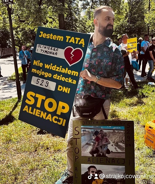 Mężczyzna z brodą trzyma transparent z napisem „Jestem TATĄ” oraz „STOP ALIENACJI” podczas protestu w parku.