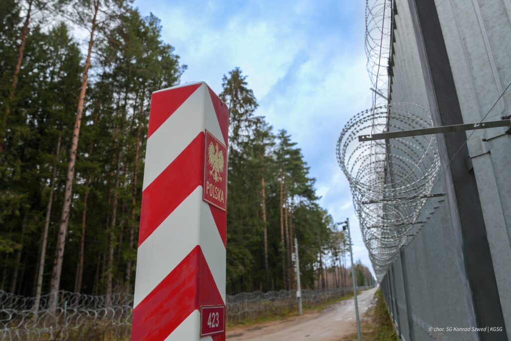 Słup graniczny z herbem Polski i numerem 423, obok ogrodzenia z drutu kolczastego, w lesie.