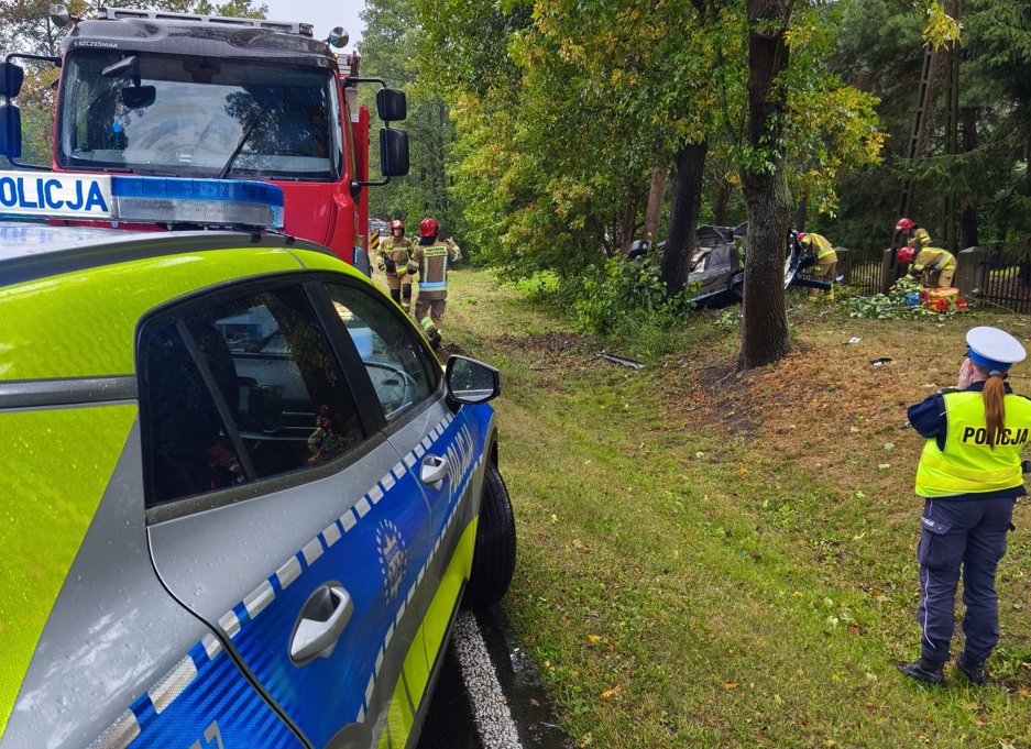 Strażacy pracują przy przewróconym samochodzie w lesie, obok radiowozu policji z włączonymi sygnałami.