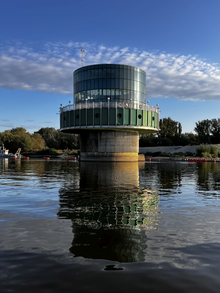 Nowoczesny budynek z przeszkloną fasadą, uniesiony nad wodą, odbija się w rzece pod błękitnym niebem.