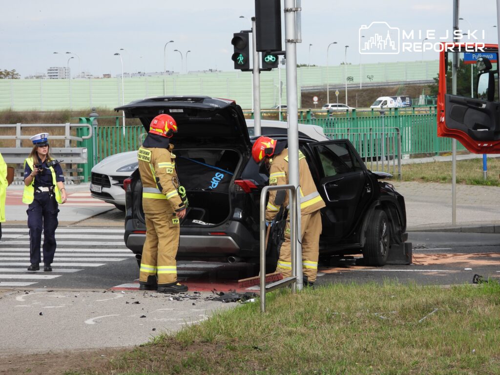 Strażacy w mundurach przeszukują wnętrze uszkodzonego pojazdu na skrzyżowaniu, obok policjantka zabezpiecza miejsce zdarzenia.
