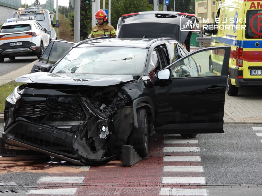 Czarny SUV z uszkodzoną przednią częścią stoi na przejściu dla pieszych, obok interweniują strażacy i karetka.