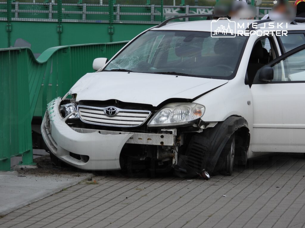 Uszkodzony biały samochód osobowy marki Toyota stojący przy zielonym ogrodzeniu na chodniku.