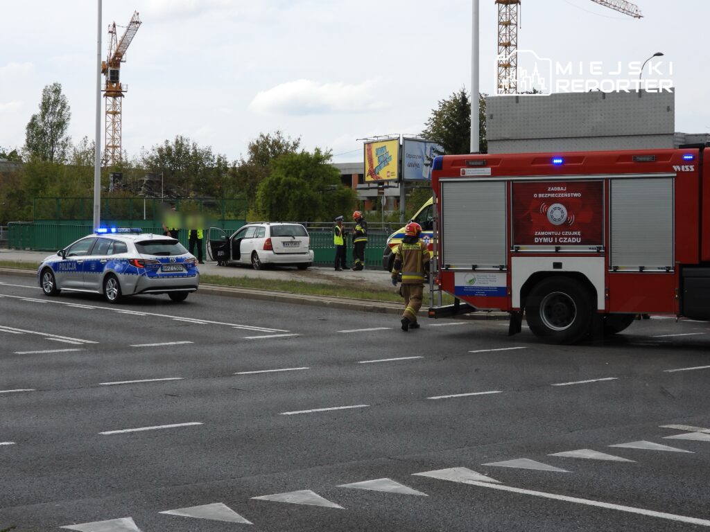 Policja i straż pożarna interweniują na drodze przy uszkodzonym pojeździe, w tle budowy i billboardy.