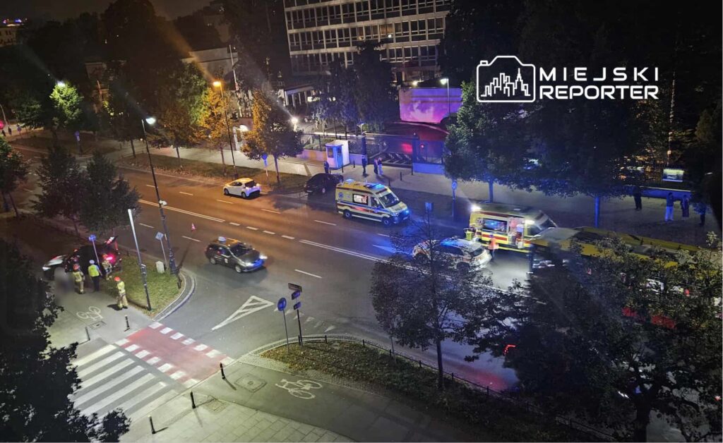 Grupa strażaków w żółtych hełmach interweniuje przy zderzeniu samochodów na ulicy, obok jadące karetki.