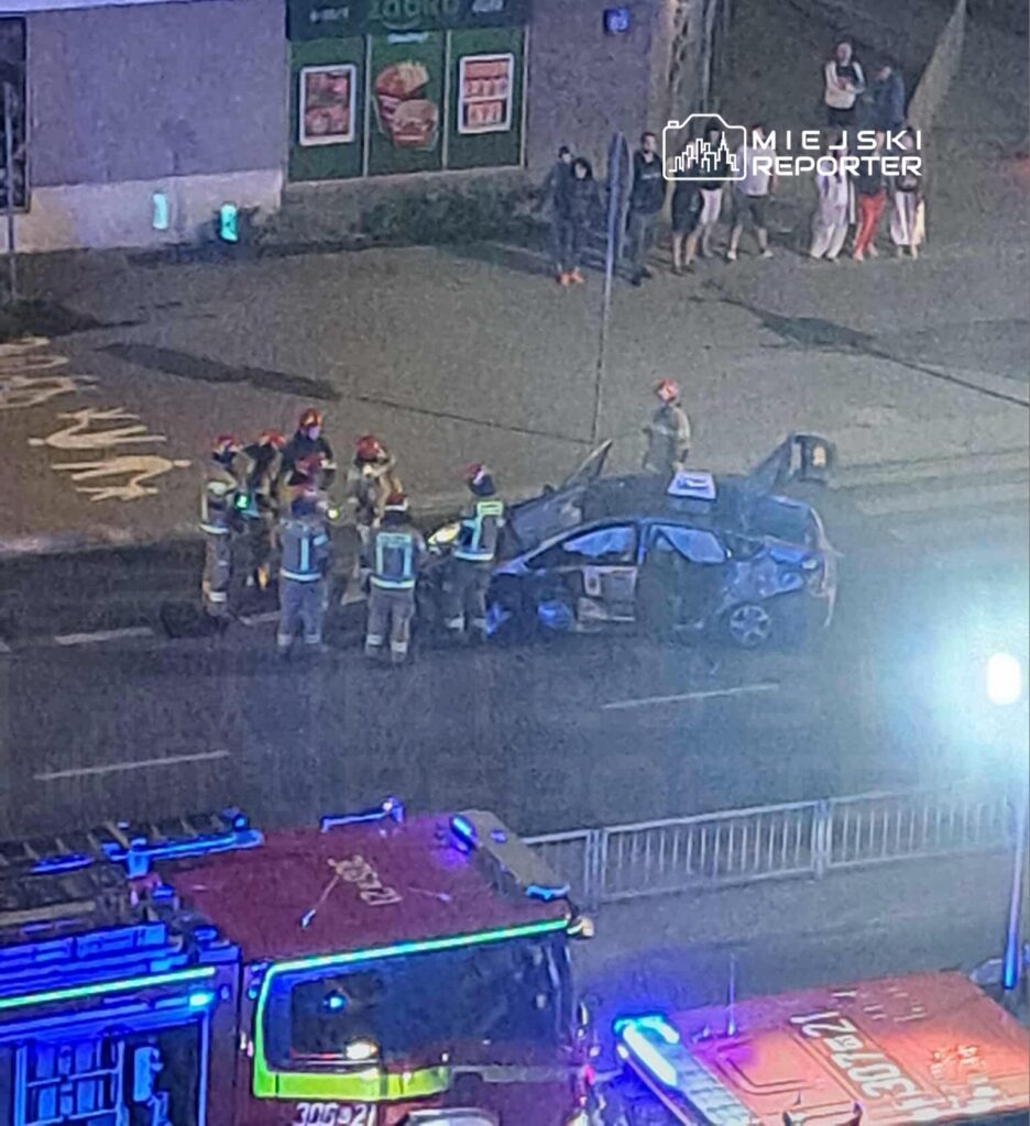 Grupa strażaków w hełmach bada uszkodzony samochód na jezdni, obok stoją wpatrzeni przechodnie.