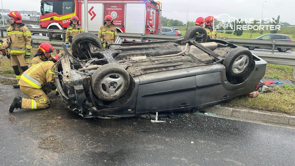 Strażacy w mundurach zabezpieczają przewrócone na dach auto na poboczu drogi, obok widać pojazd strażacki.