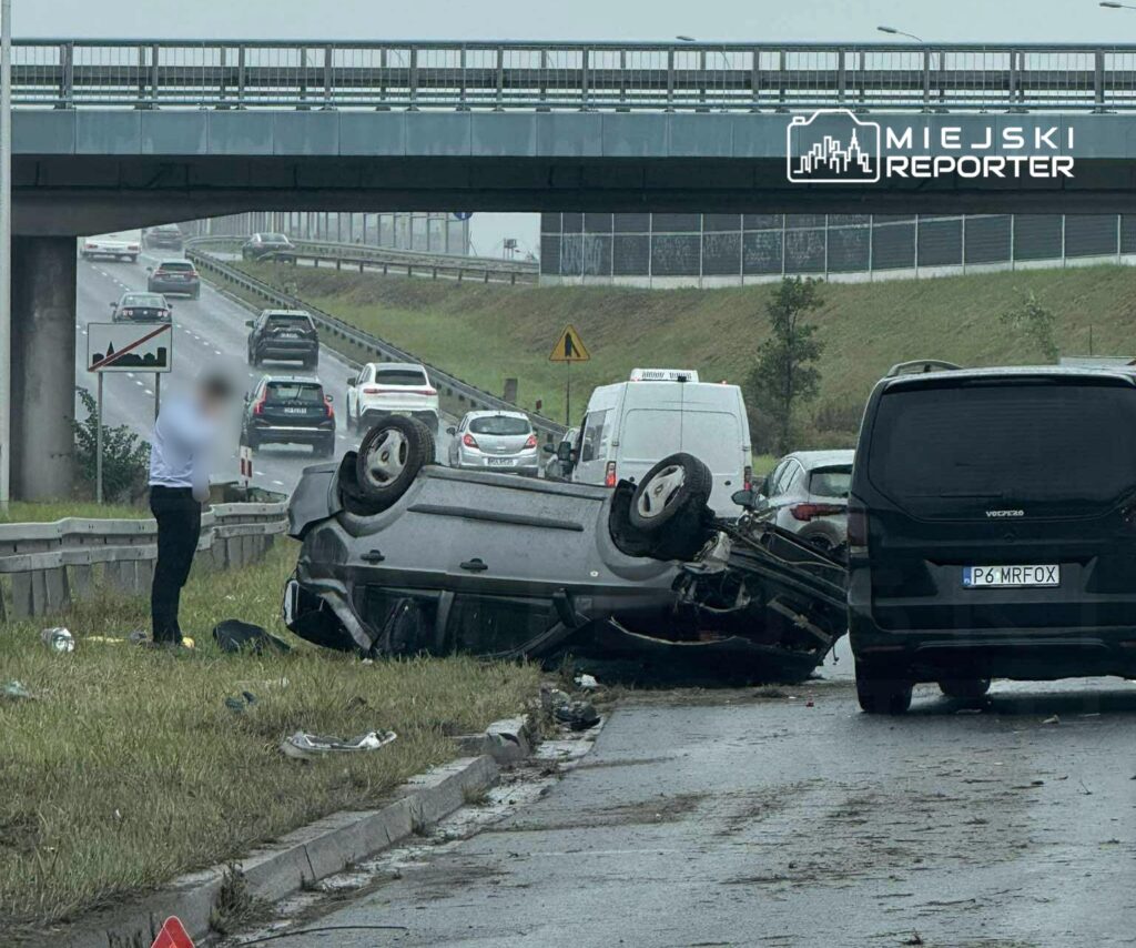 Na drodze leży przewrócone auto, a obok stoi mężczyzna w białej koszuli. W tle widać inne pojazdy w ruchu.