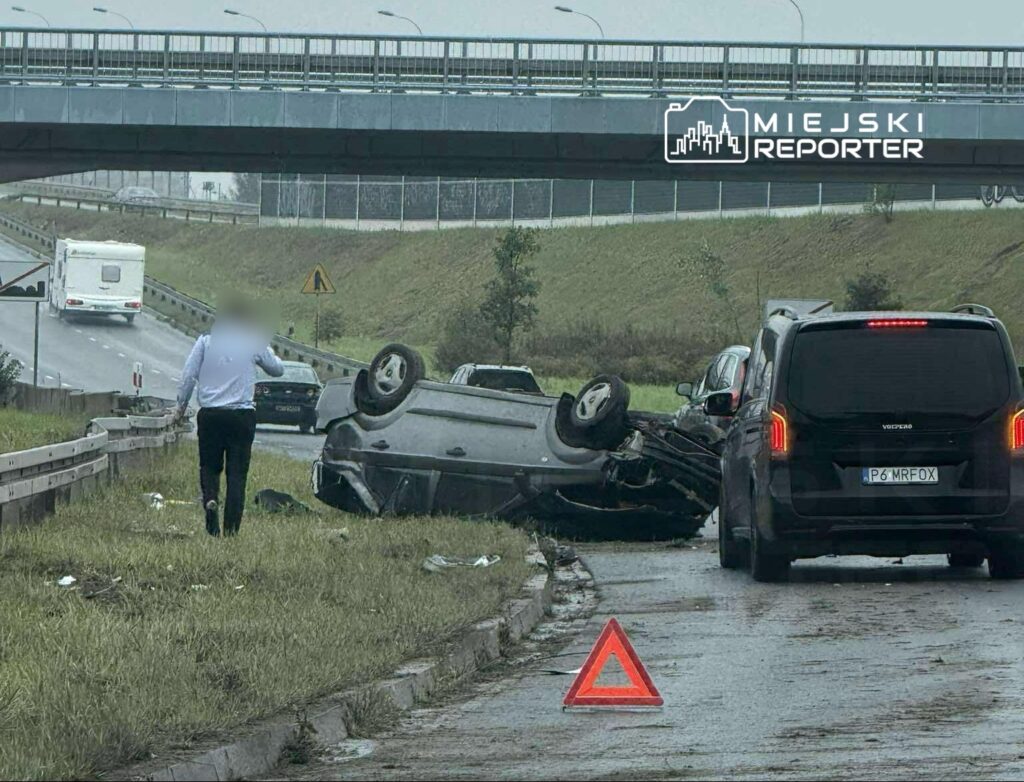 Na drodze leży przewrócony samochód, obok mężczyzna w białej koszuli idzie w kierunku pojazdu, a w tle widać inne samochody.