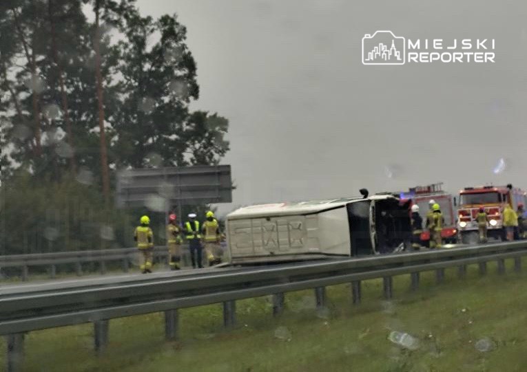 Zespół ratunkowy w żółtych hełmach pracuje przy przewróconym pojeździe na poboczu drogi, w tle widać straż pożarną.