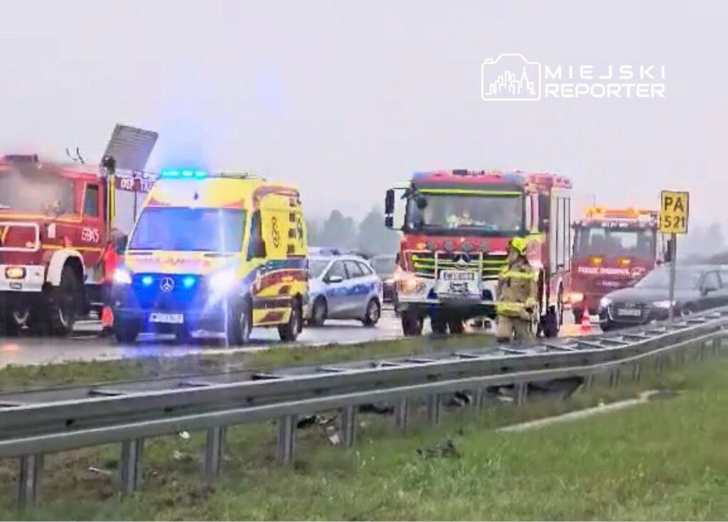 Karetka i samochody straży pożarnej interweniują na drodze, obok stojących pojazdów w deszczowej pogodzie.