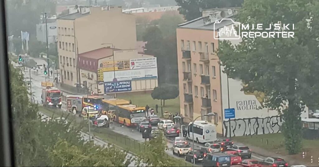 Na ulicy stoją pojazdy ratunkowe, a obok grupka ludzi obserwuje sytuację w deszczowej pogodzie.