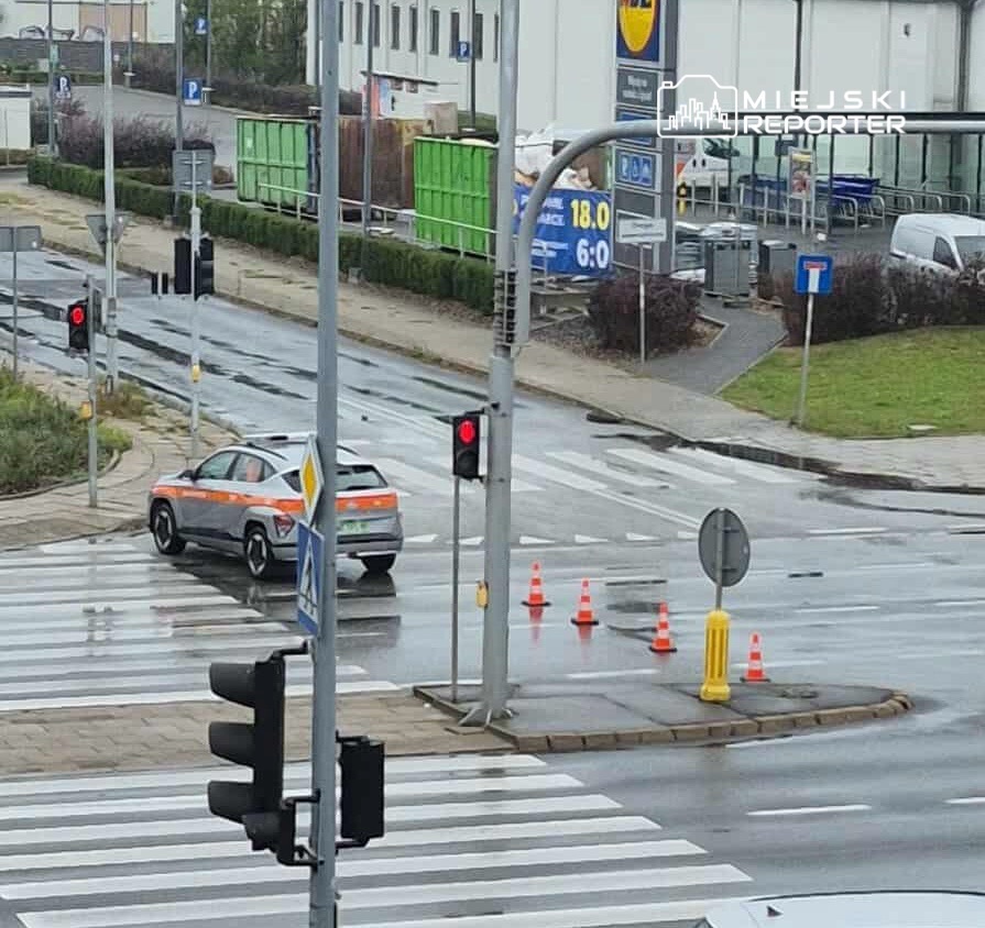 Samochód służbowy z pomarańczowym oznakowaniem skręca w lewo na mokrej ulicy z czerwonymi sygnalizatorami.